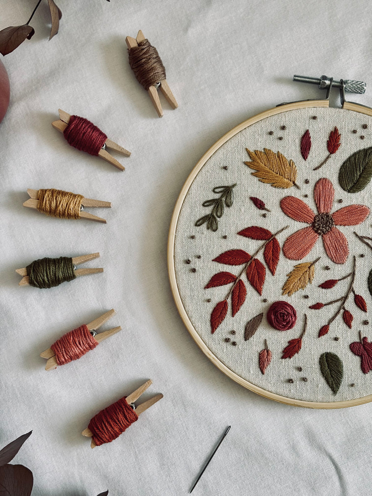 Detalle de flor bordada en tonos rojizos y hojas otoñales en bastidor, junto a hilos de bordado
