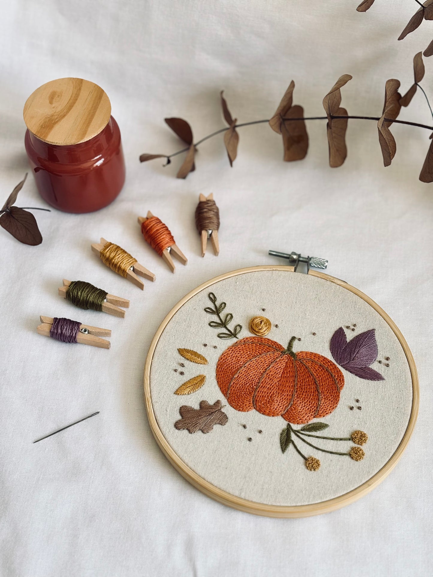 Bordado a mano terminado con diseño de calabaza naranja, hojas doradas y moradas, y flores silvestres en tonos otoñales, con hilos DMC organizados en pinzas